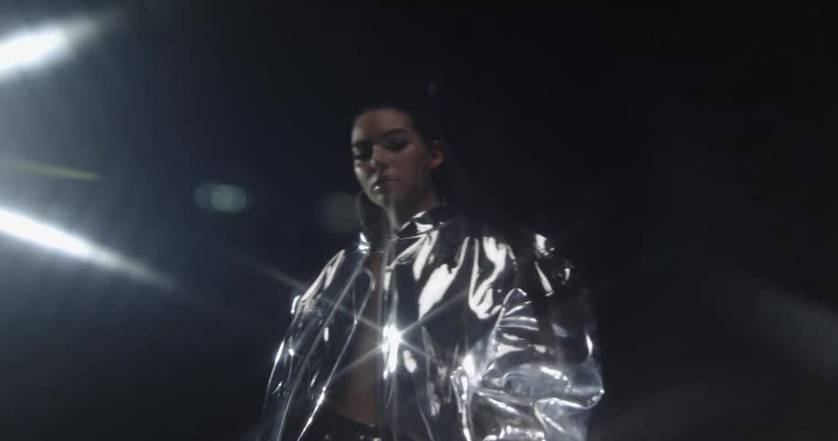 A woman dances under a bright light in a silver jacket and matching skirt
