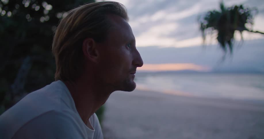 A man is sitting on a beach and looking around as waves roll over the sand