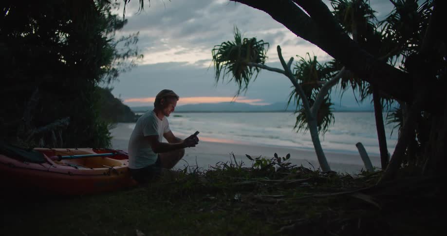 A man is sitting on a beach using his cell phone