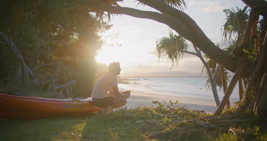A man carries a kayak to the edge of a beach and sets it down to take pictures with his cell phone