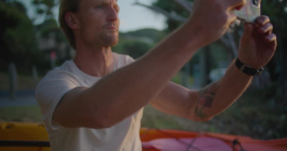 Blonde man with white t shirt and black shorts squatting down next to yellow and red boat takes photos on smartphone