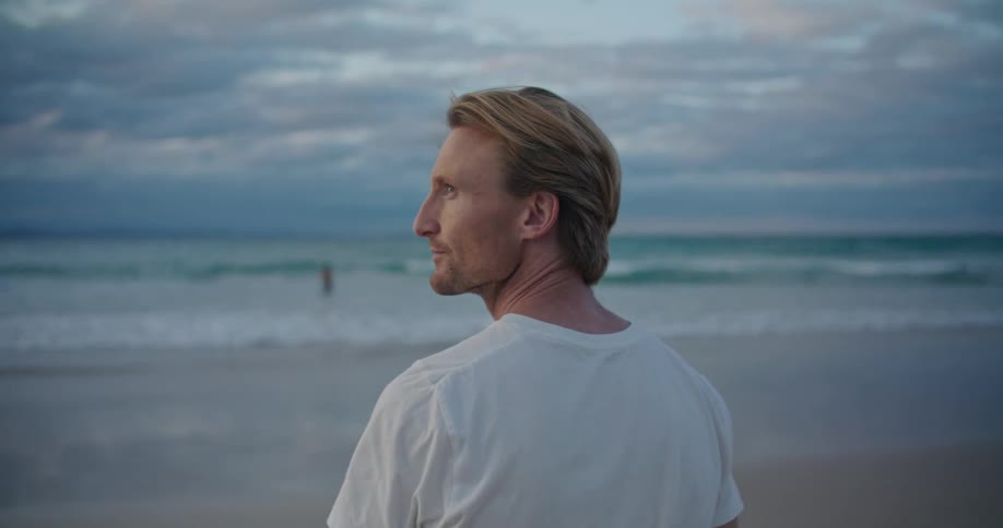 A man wearing a white tshirt is standing on a beach watching the waves
