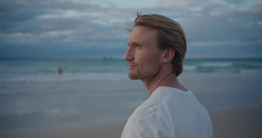 Man stands on beach looking toward ocean as another person walks out of water