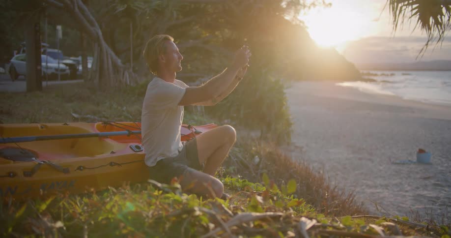 A man kneeling in front of his kayak uses a smartphone to take a photograph of a beach