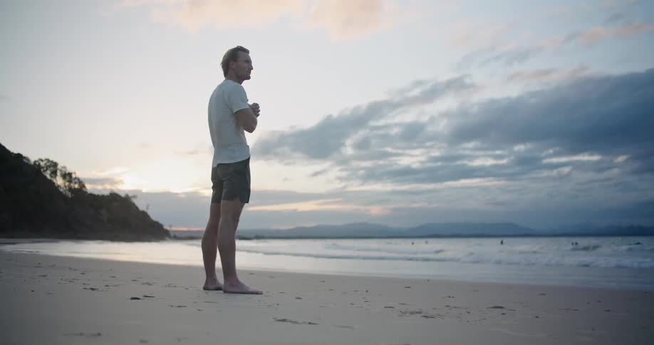 A man stands on the beach sand looking at the waves and stretching his legs