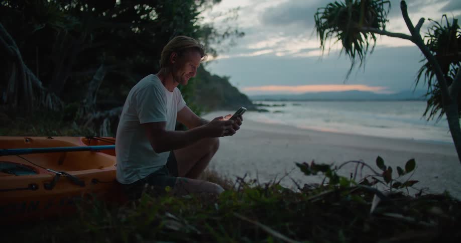 A man texts on his phone while sitting next to his kayak on the beach