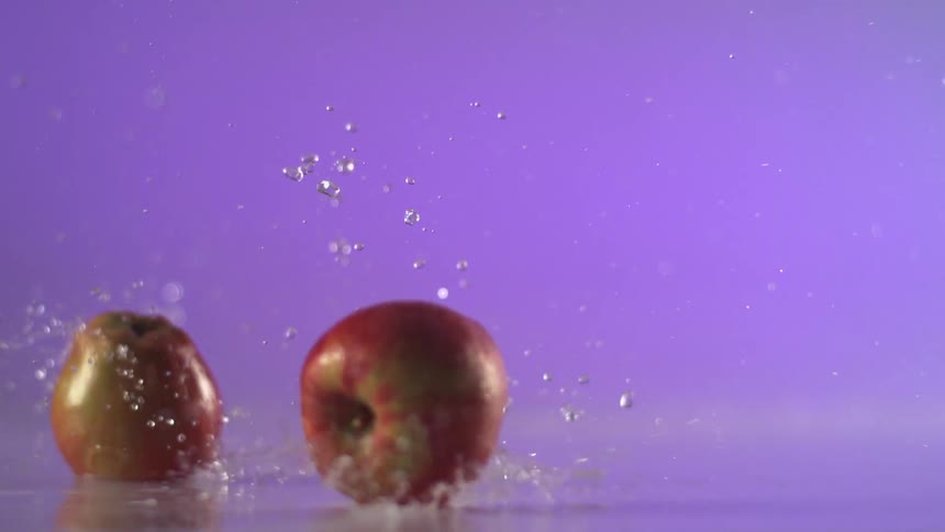 Two apples are filmed falling and bouncing atop a light layer of water in a studio setting