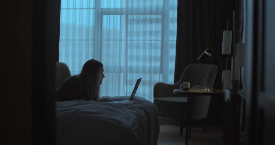 A woman in a room laying on a bed and using a laptop with a wall and window in the background