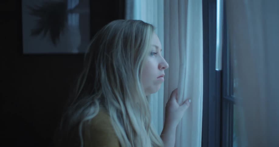 A woman in a room looking out of a window with a wall and picture frame in the background