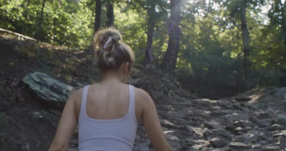 A woman is walking on a trail in the woods with the sun shining brightly upon her