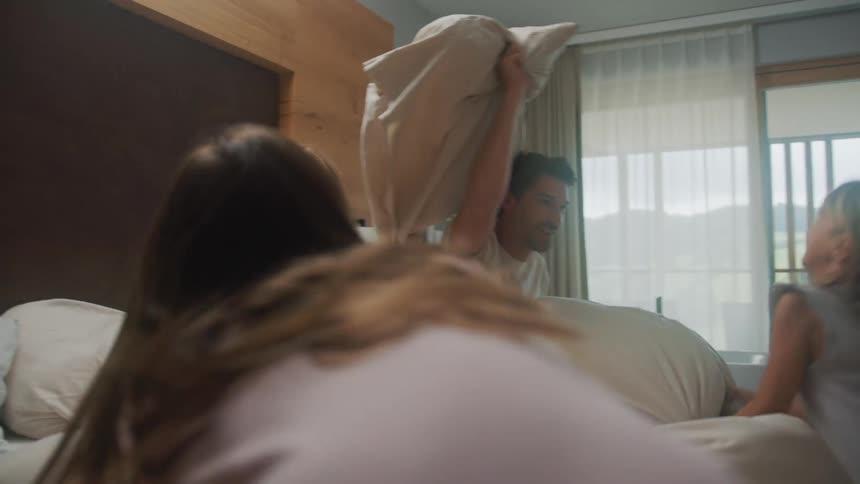 A mother and father laugh as they have a fun family pillow fight in bed with their two daughters