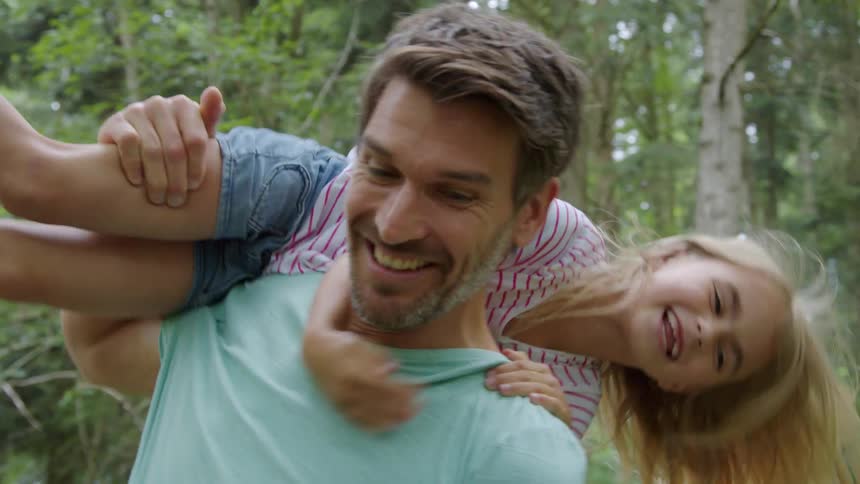 Father and daughter are playing in the woods as he swings her around on his shoulders