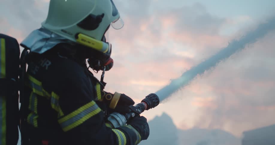 A firefighter with a helmet spraying a firehose with the sky and clouds in the background