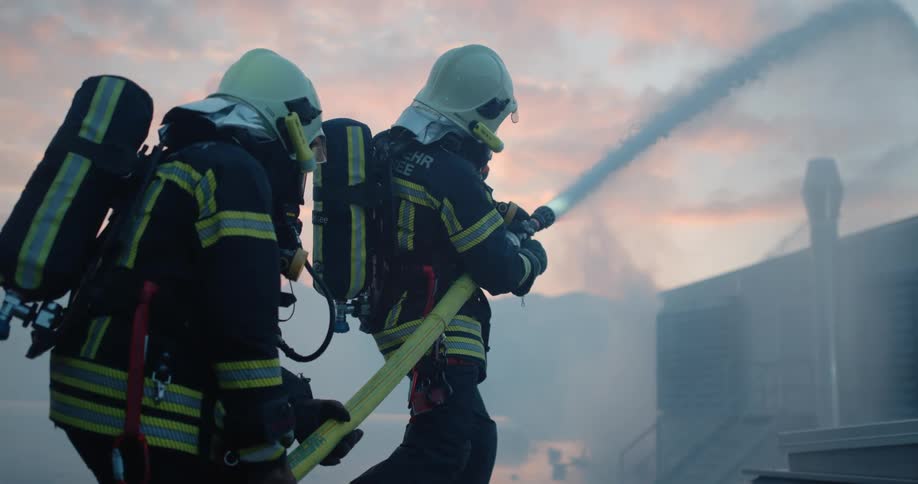 Two firefighters hold a hose spraying water onto a building