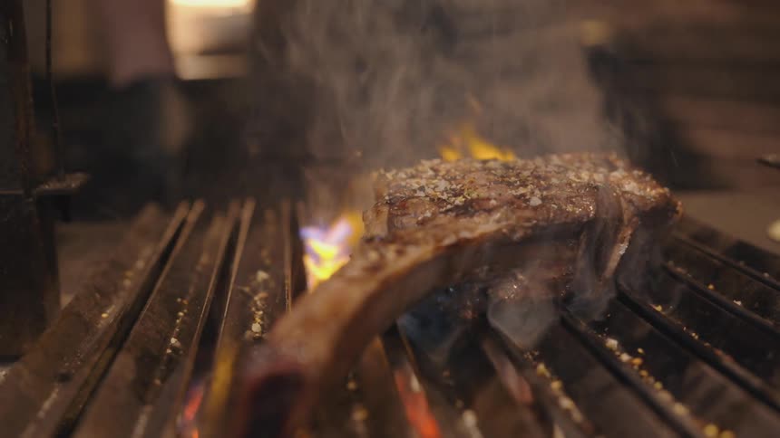 A chefs gloved hand sprinkles salt and seasoning onto a thick prime rib chop on a flaming grill