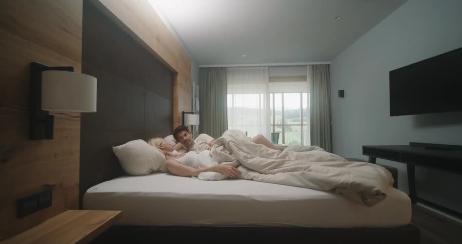 A couple lays in bed talking with sheers pulled back on the balcony doors
