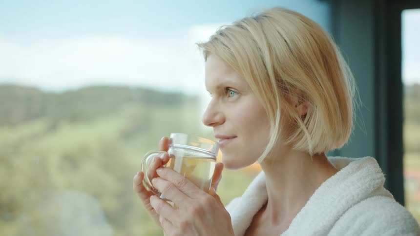 Young short haired blonde woman drinking tea while looking out the window at home