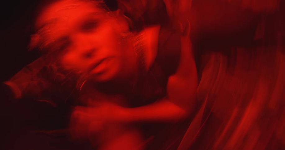 A young woman in a metallic jumpsuit rolls around in a dark room with red lighting