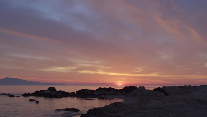 Sun brightens spot on horizon turning sky red and purple as gentle waves roll against rocky shore