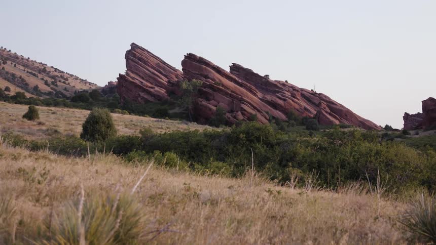 Reddish outcropping rise from grassy fields near scrub covered slope under clear light blue sky