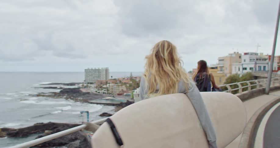 Two blonde friends carrying surfboards walks along highway pavement in coastal town