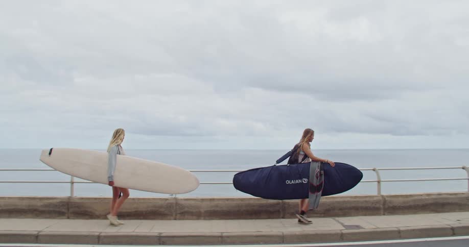 Two teenage girls walk along the boardwalk carrying their surf boards