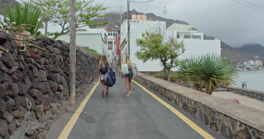Two women carrying several bags walk down a road into a town