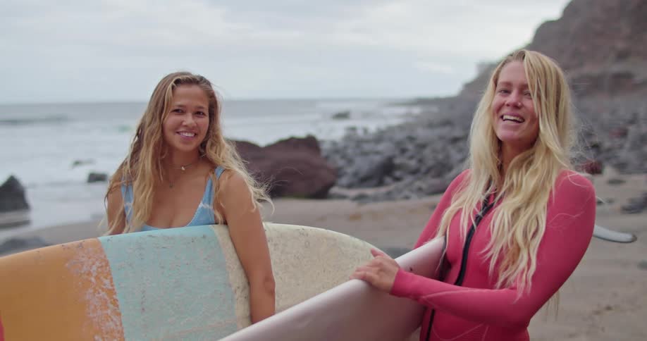 Two blonde female surfers hold surfboards while standing on beach and smiling