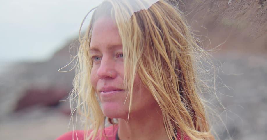 Blonde woman with piercing and pink wetsuit smiles and looks around on the coast