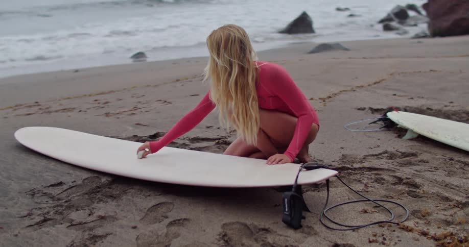 A young woman sits on the beach and waxes her surfboard