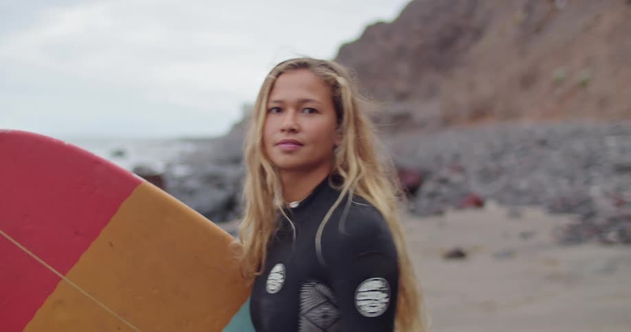 A surfer poses on the beach with a surfboard under her arm before walking toward the ocean