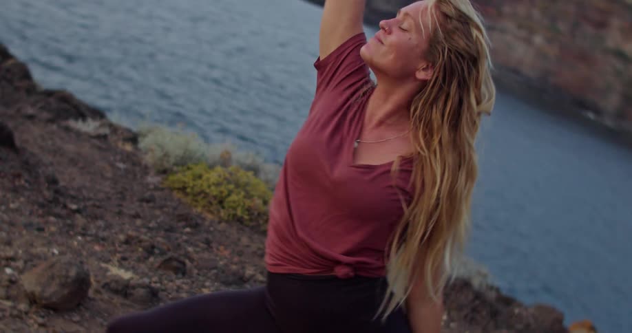Blonde woman moves through yoga poses on shore across water from cliff side