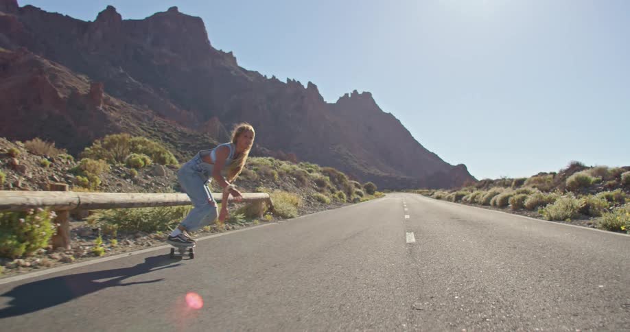 Blonde woman with strappy denim jumpsuit rides on skateboard on highway