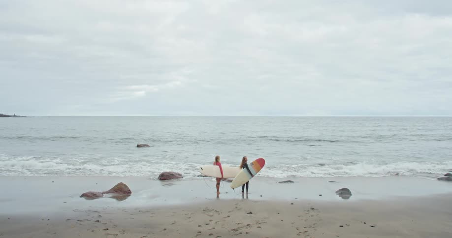 Two women with surfboards standing on a beach with the ocean the sky and clouds in the background