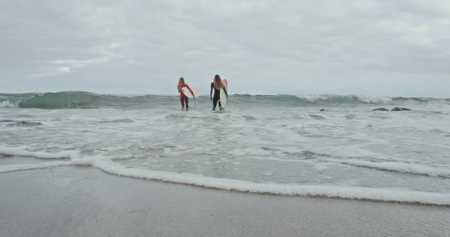 Two women carry surfboards under their arms as they walk into the ocean