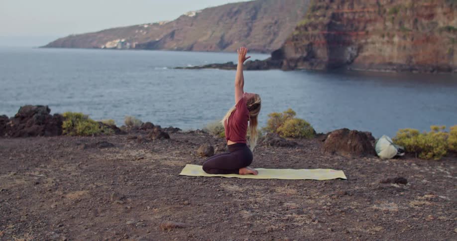 Blonde woman performs yoga on yellow mat on shore across water from cliff