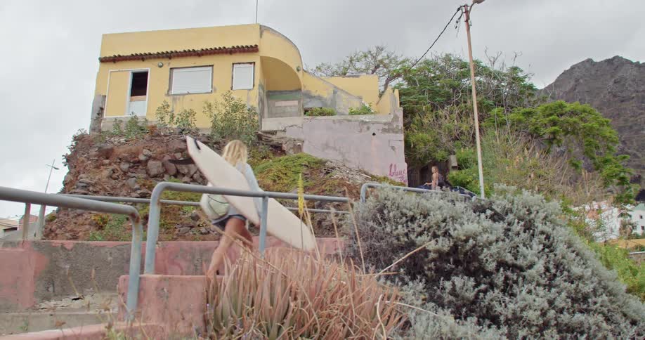A woman with a surfboard walking towards a building with the sky and clouds in the background