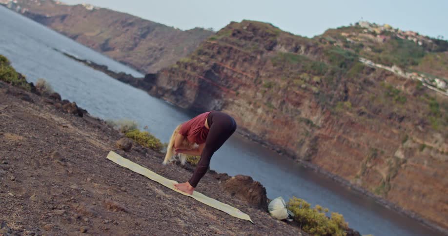 A woman bends down while doing yoga alongside a lake in a canyon