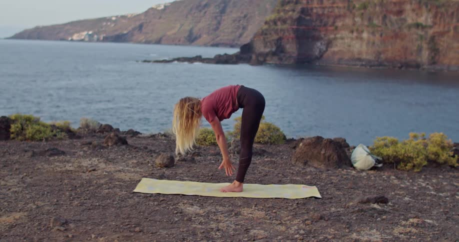 Blonde woman moves through yoga poses on shore across from cliff