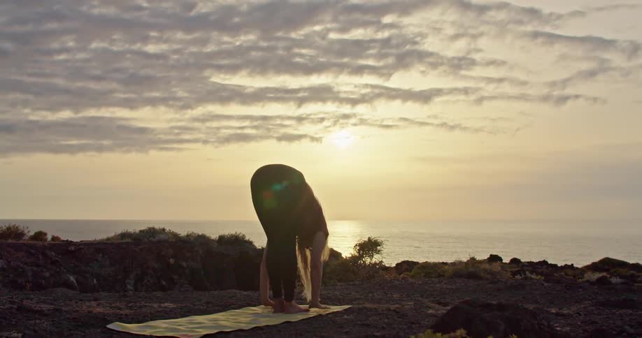 Barefoot blonde woman moves through yoga posses on yellow yoga mat set on shore with ocean reflecting sun in background