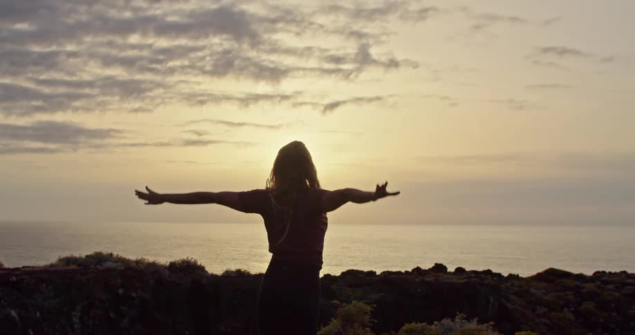 A woman is doing yoga poses on a hilltop overlooking the ocean at sunset