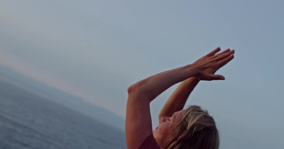 A woman stretches her arms up and back down while doing yoga on a cliff over the ocean