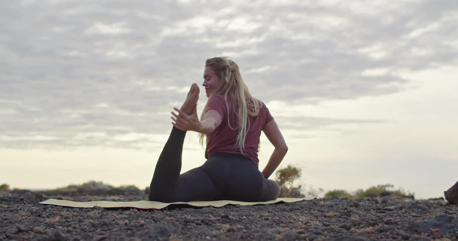 A woman grabs her foot from behind while doing yoga outdoors on a cliff