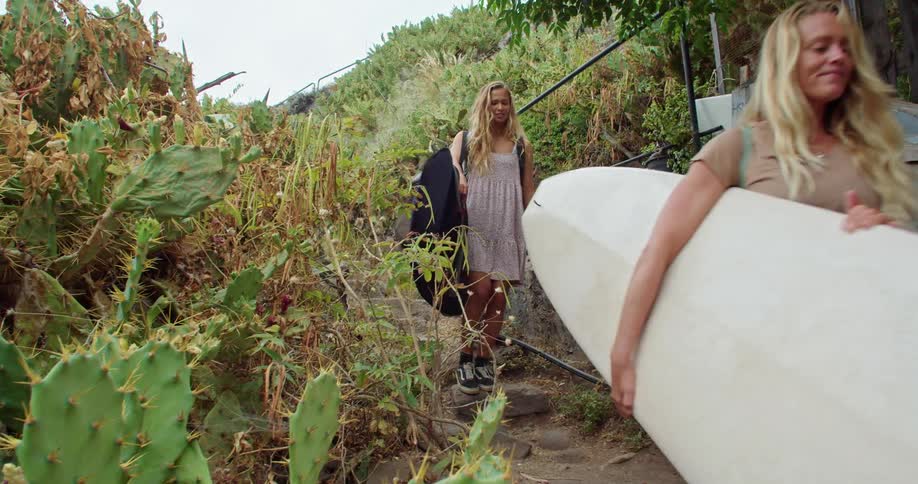 Two blonde women walk down stone steps carrying surfboards and smiling
