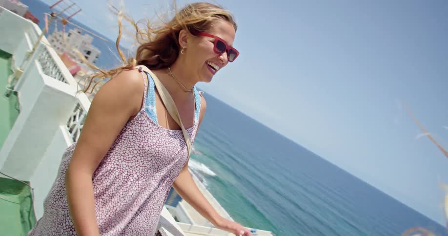 Two blonde women with sunglasses talk with each other laughing on green hotel terrace overlooking town and beach