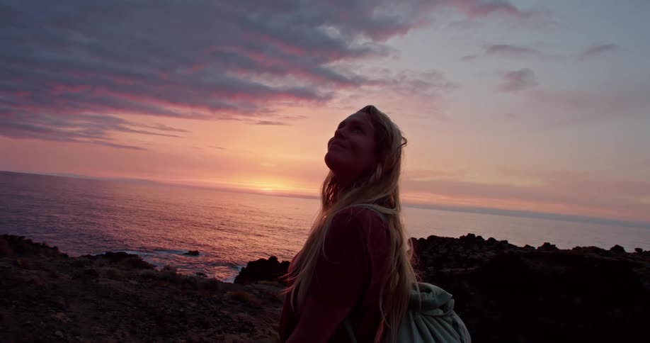 Blonde woman stands on shores looking around at sky as glimmer on sun on horizon turns sky red
