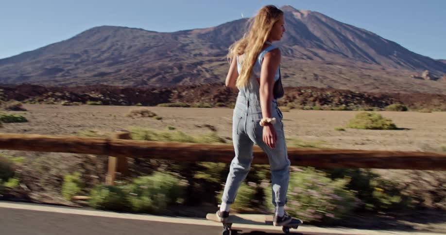Blonde woman with strappy denim jumpsuit rides on skateboard on highway