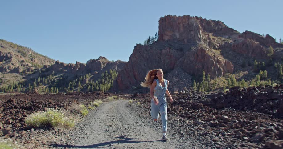 Blonde woman with strappy denim jumpsuit runs along dirt roadsmiling
