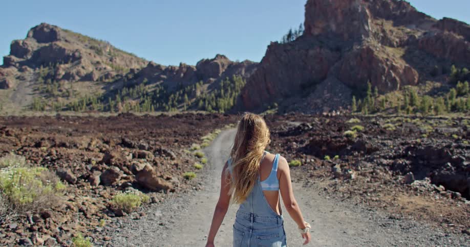 A young woman is walking on a dry dirt road towards large boulders on sunny day