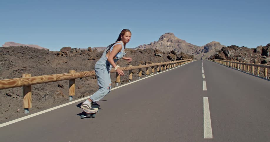 Woman rides skateboard across long bridge spanning rough terrain with rocky mountains and mesa in background